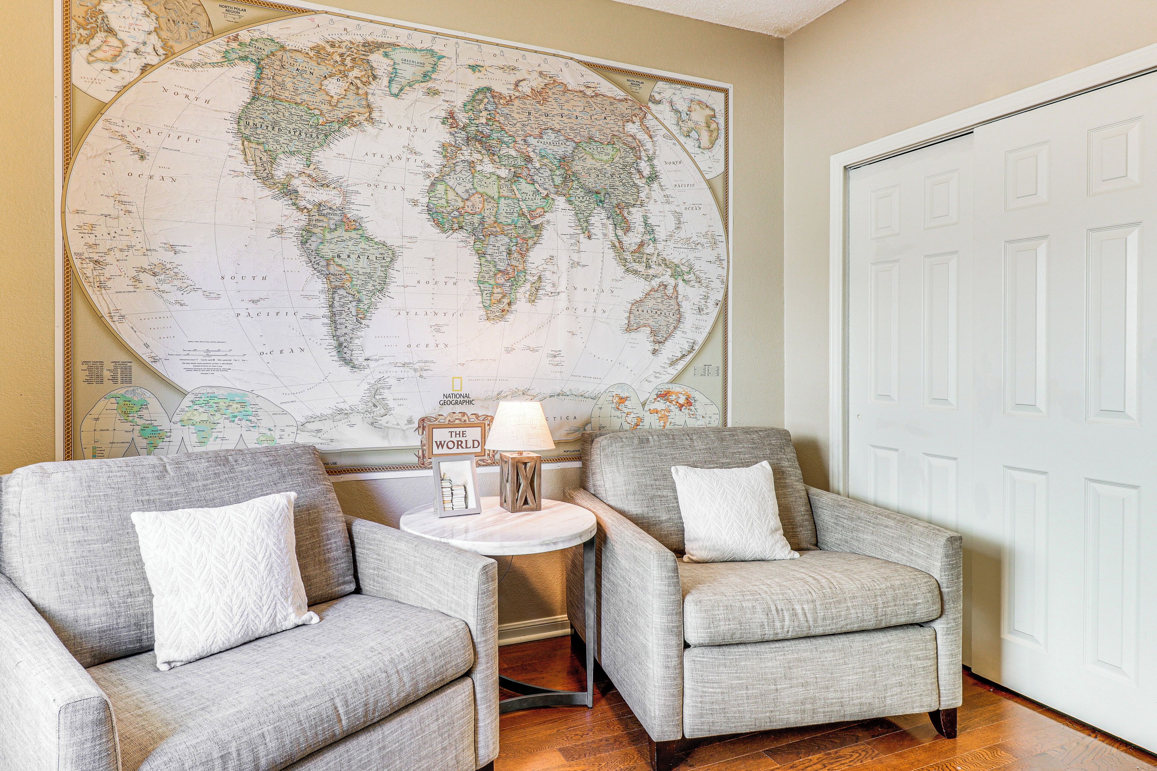 Une pièce avec deux fauteuils gris, une table ronde avec une lampe et une grande carte du monde accrochée au mur.