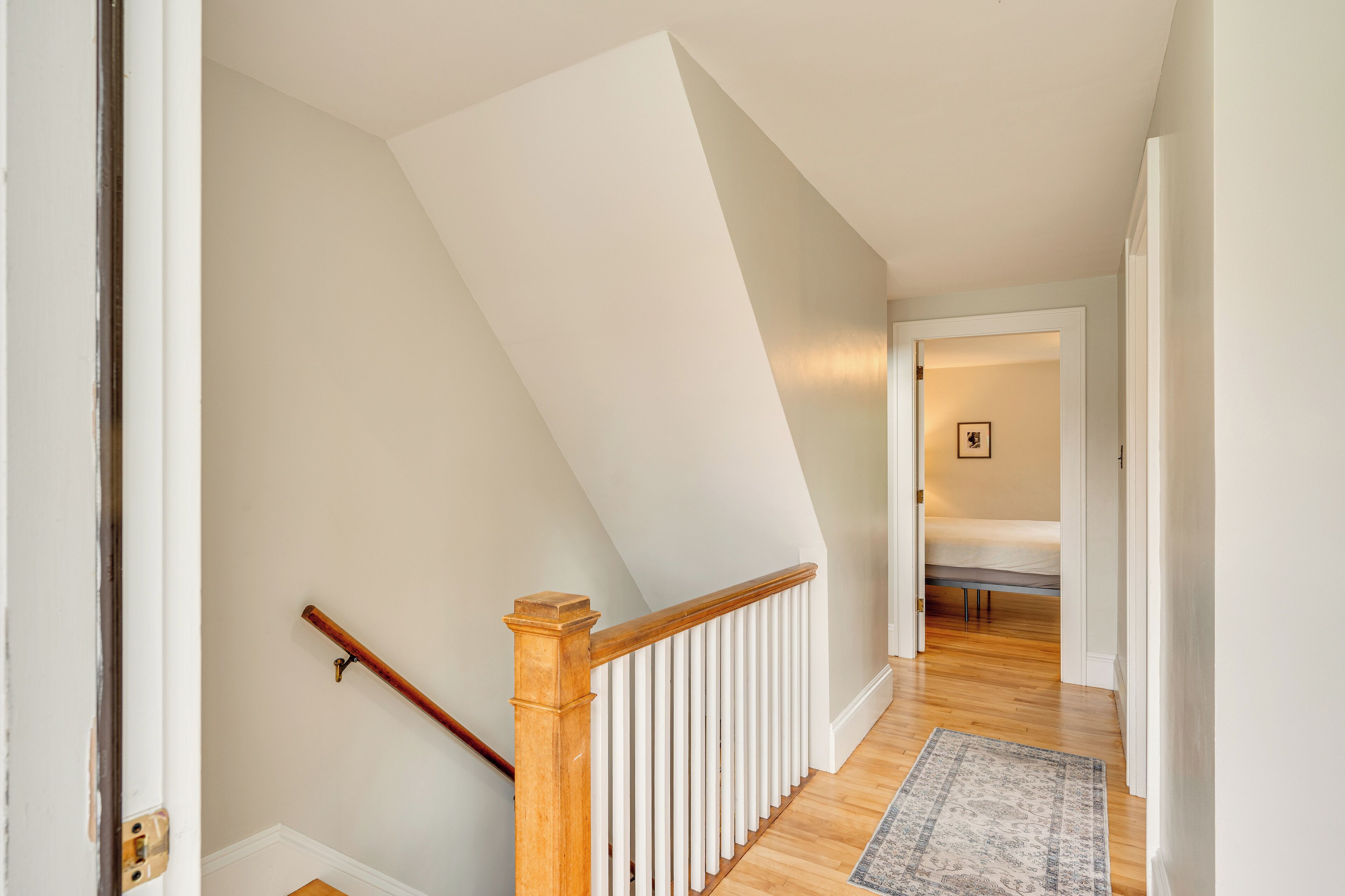 A staircase with a wooden handrail and white balusters, leading to a bedroom with a bed and a framed picture on the wall.
