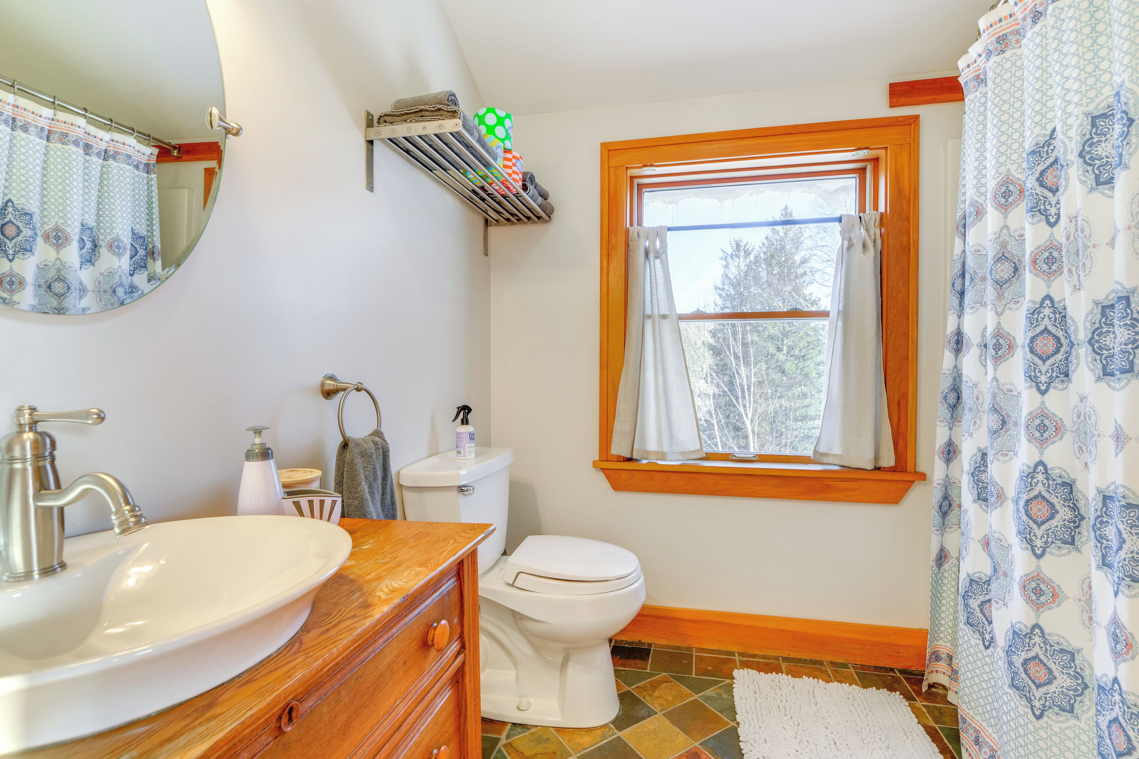 A bathroom with a white sink, wooden vanity, toilet, and a window with a view of trees.