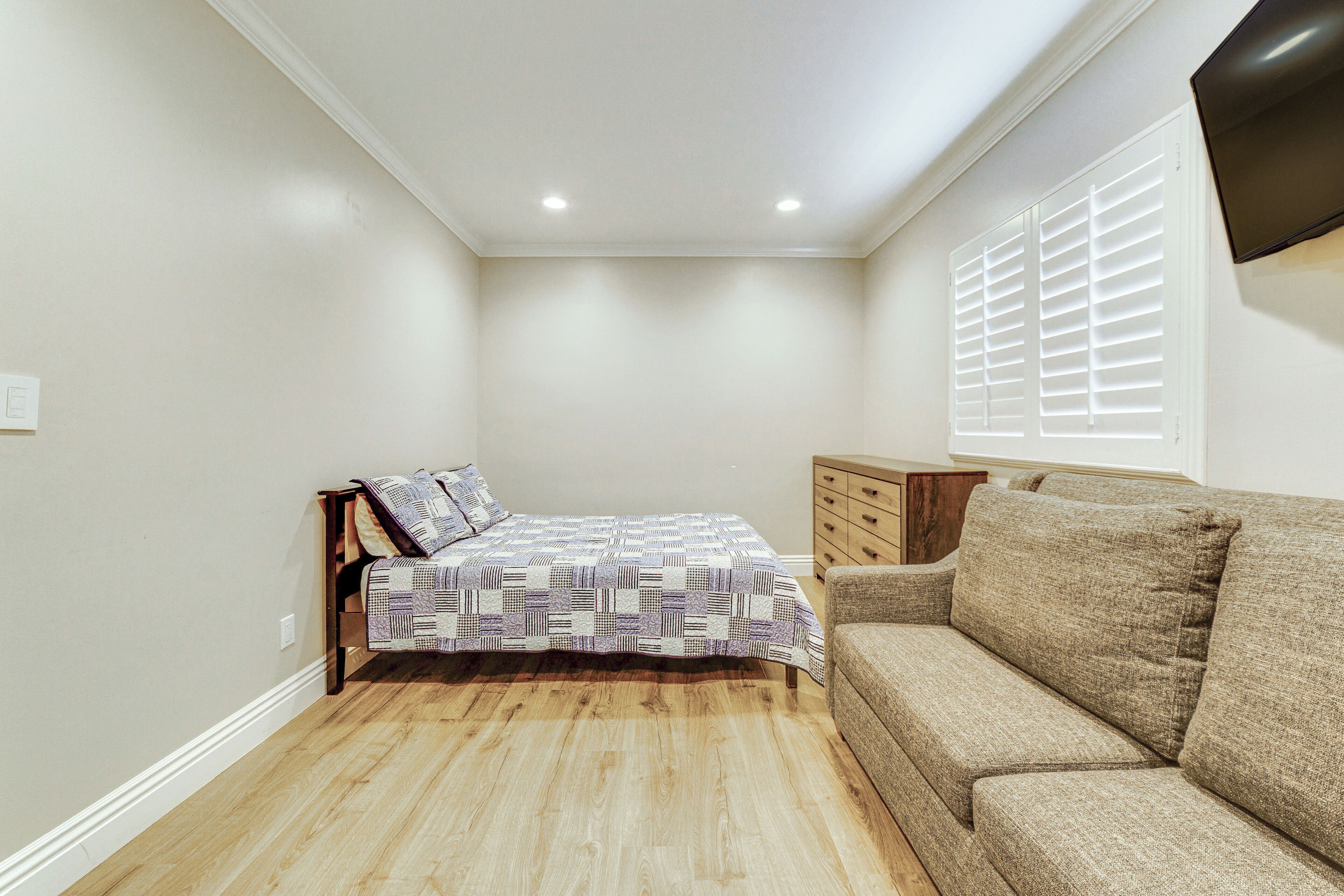 A bedroom with a bed, a sofa, a wooden dresser, and a window with blinds.
