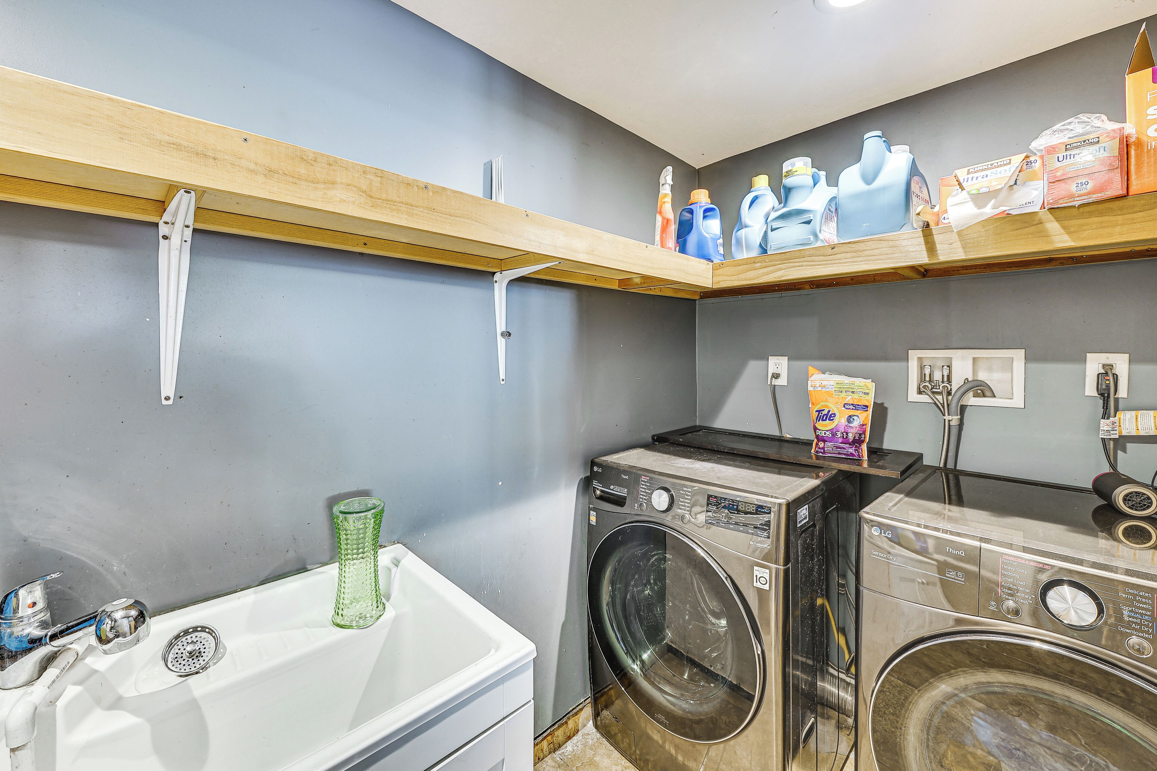 A laundry area with a washing machine, a sink, and shelves with detergent bottles.