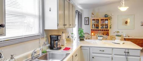 A kitchen with white cabinets, a double sink, a coffee maker, and a window with blinds.