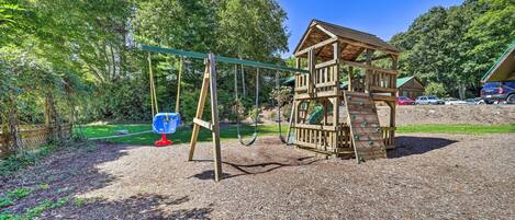 A wooden play structure with a slide and swings, surrounded by trees and a clear sky.