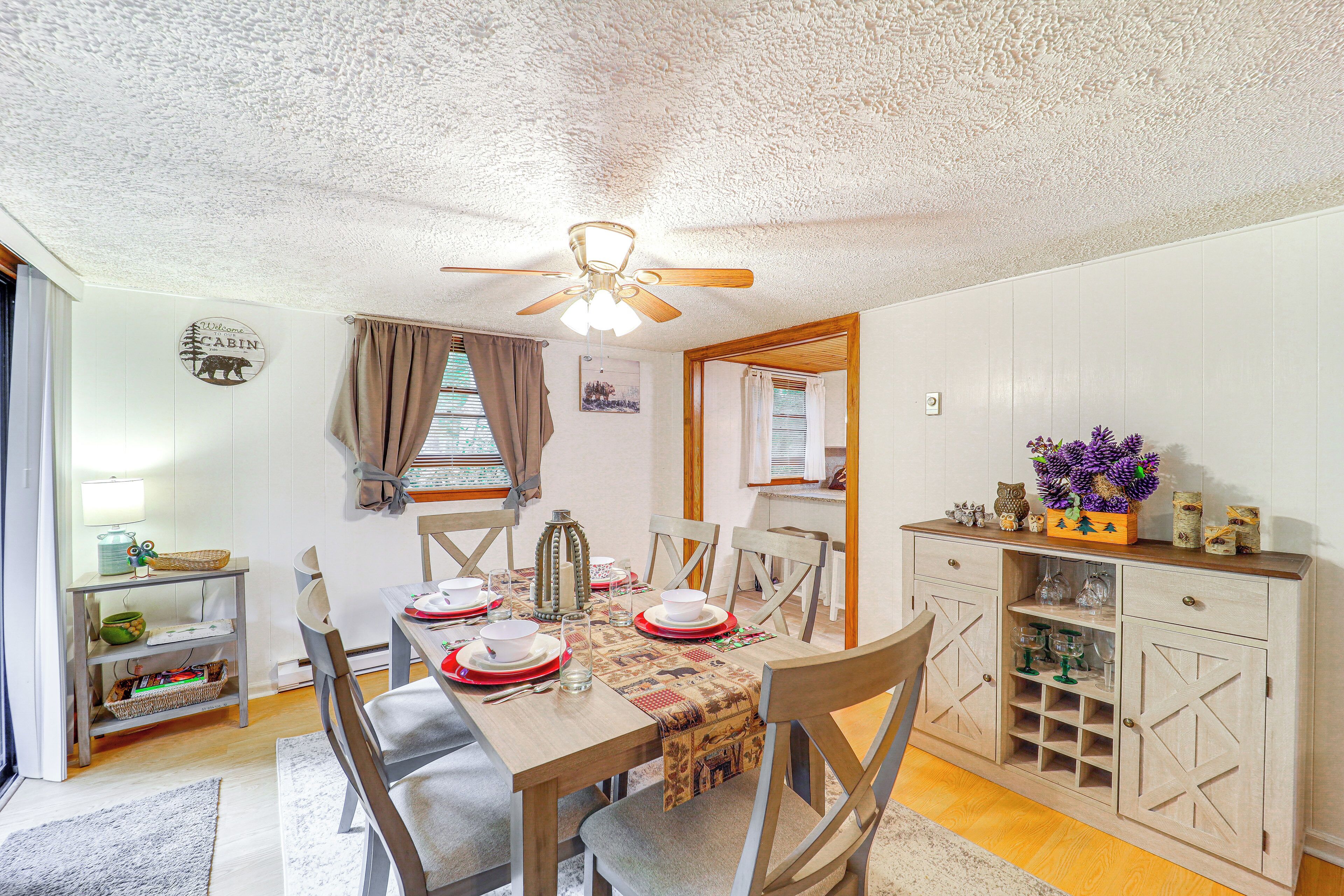 A dining area with a wooden table set for four, a ceiling fan, and a cabinet with glassware.