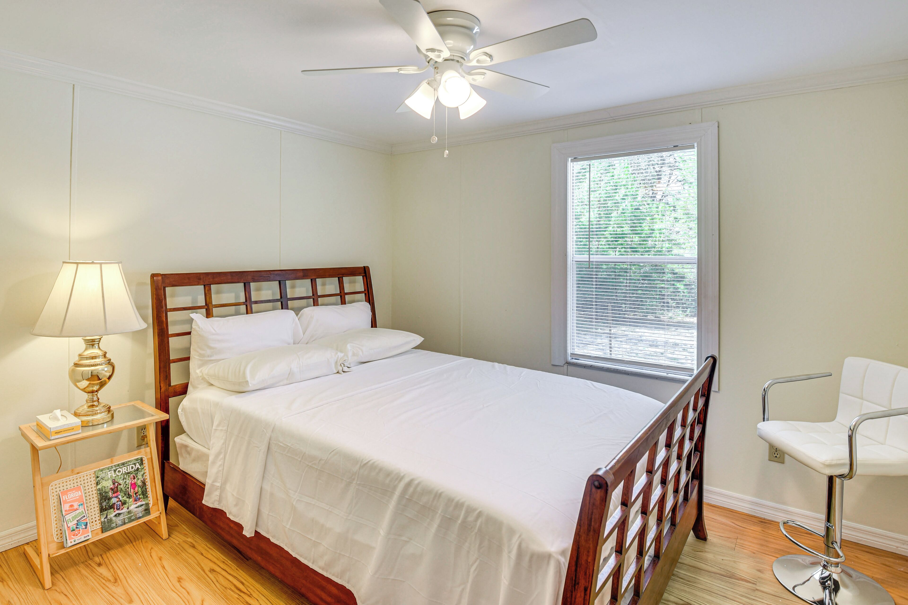 A neatly made bed with white linens, a wooden headboard, a bedside table with a lamp, and a window with blinds.