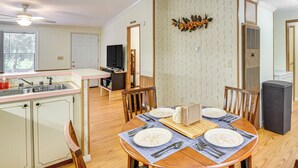 Un comedor con una mesa de madera dispuesta para cuatro, una cocina con un fregadero y armarios, y un televisor al fondo.