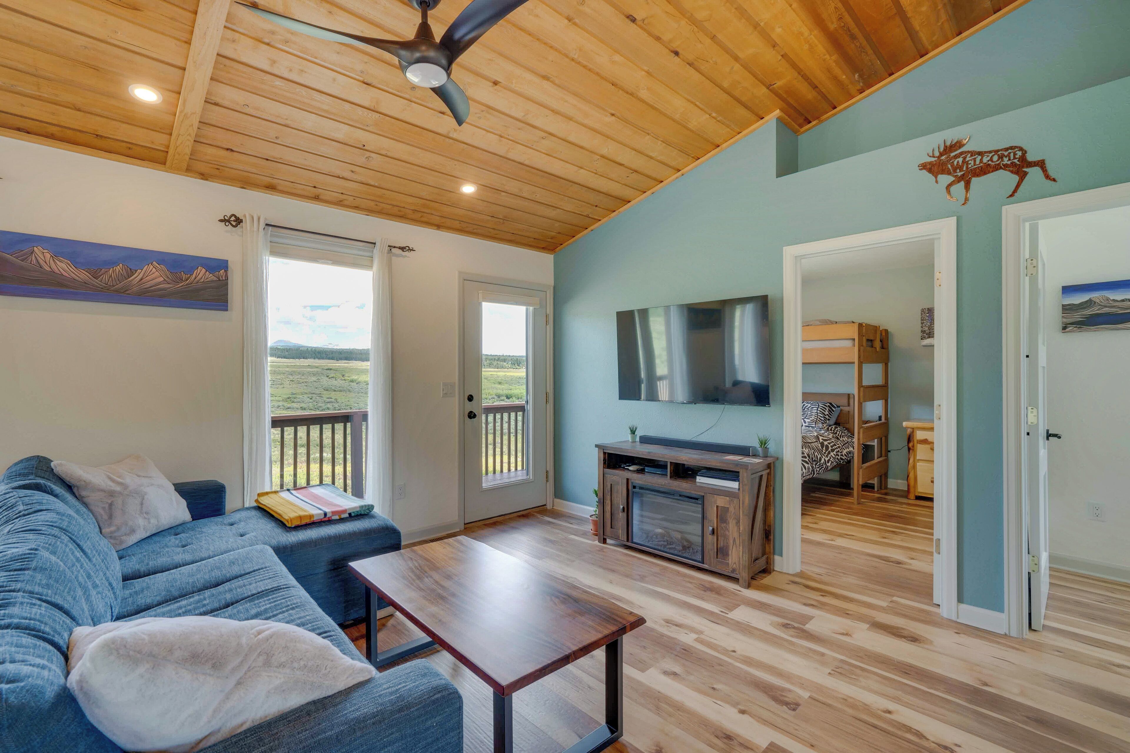 A spacious living room with a blue sofa, wooden coffee table, and a television. There is a wooden ceiling, a door leading to another room, and a view of the outdoors through a glass door.