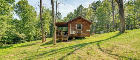 A wooden cabin with a deck, surrounded by trees and grassy terrain.