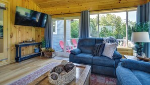 A cozy living room with a blue sofa, a flat-screen TV, wooden walls, and a large window with curtains. - Mountain-view Maine Cabin: Ski, Hike & Bike! (Strong)