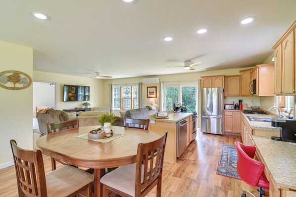 A spacious kitchen with wooden cabinets, a dining area with a wooden table and chairs, and a view of the outdoors through large windows. - Mountain-view Maine Cabin: Ski, Hike & Bike! (Strong)