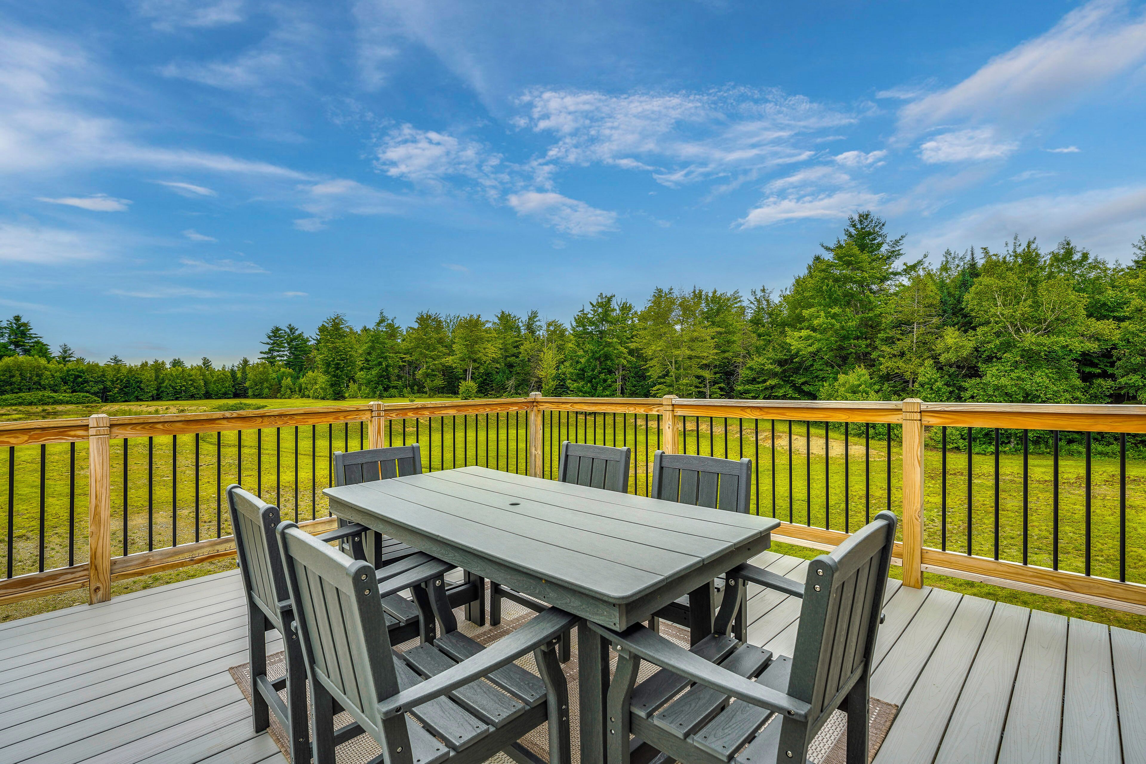A deck with a table and chairs, surrounded by a wooden railing, overlooking a grassy area with trees.