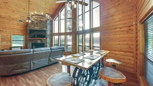 A log cabin interior with a stone fireplace, wooden ceiling, large windows, a sofa, a dining table, and stools. - Lakefront Gladewater Retreat w/ Dock + Hot Tub! (Gladewater)