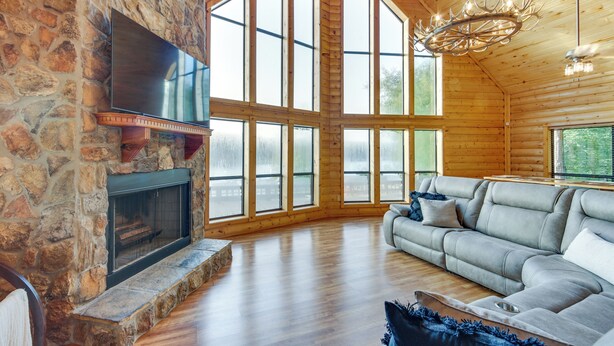 A spacious living room with a stone fireplace, large windows, a grey sofa, and a chandelier. - Lakefront Gladewater Retreat w/ Dock + Hot Tub! (Gladewater)