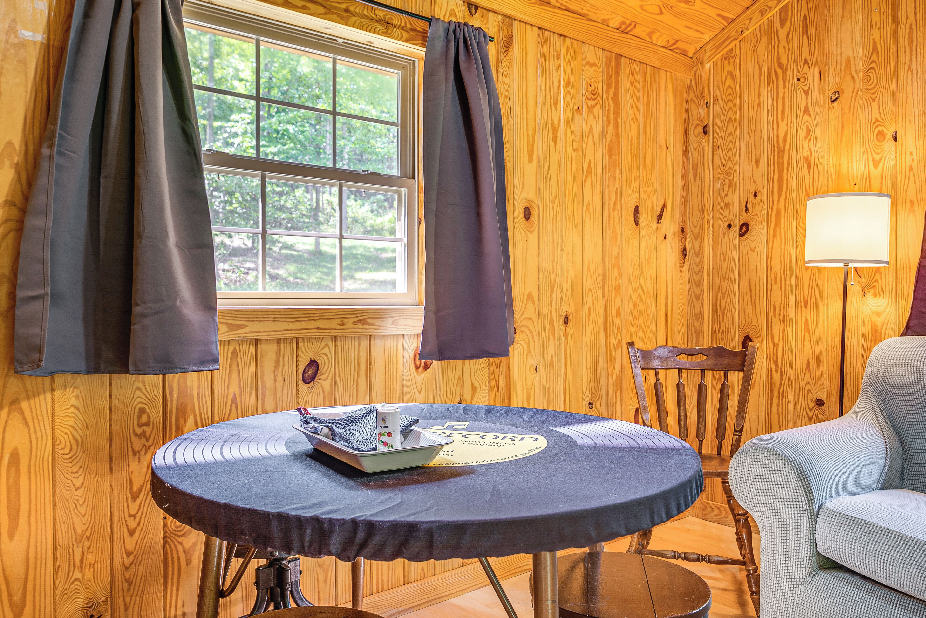 A wooden room with a round table, a chair, a lamp, and a window with curtains.