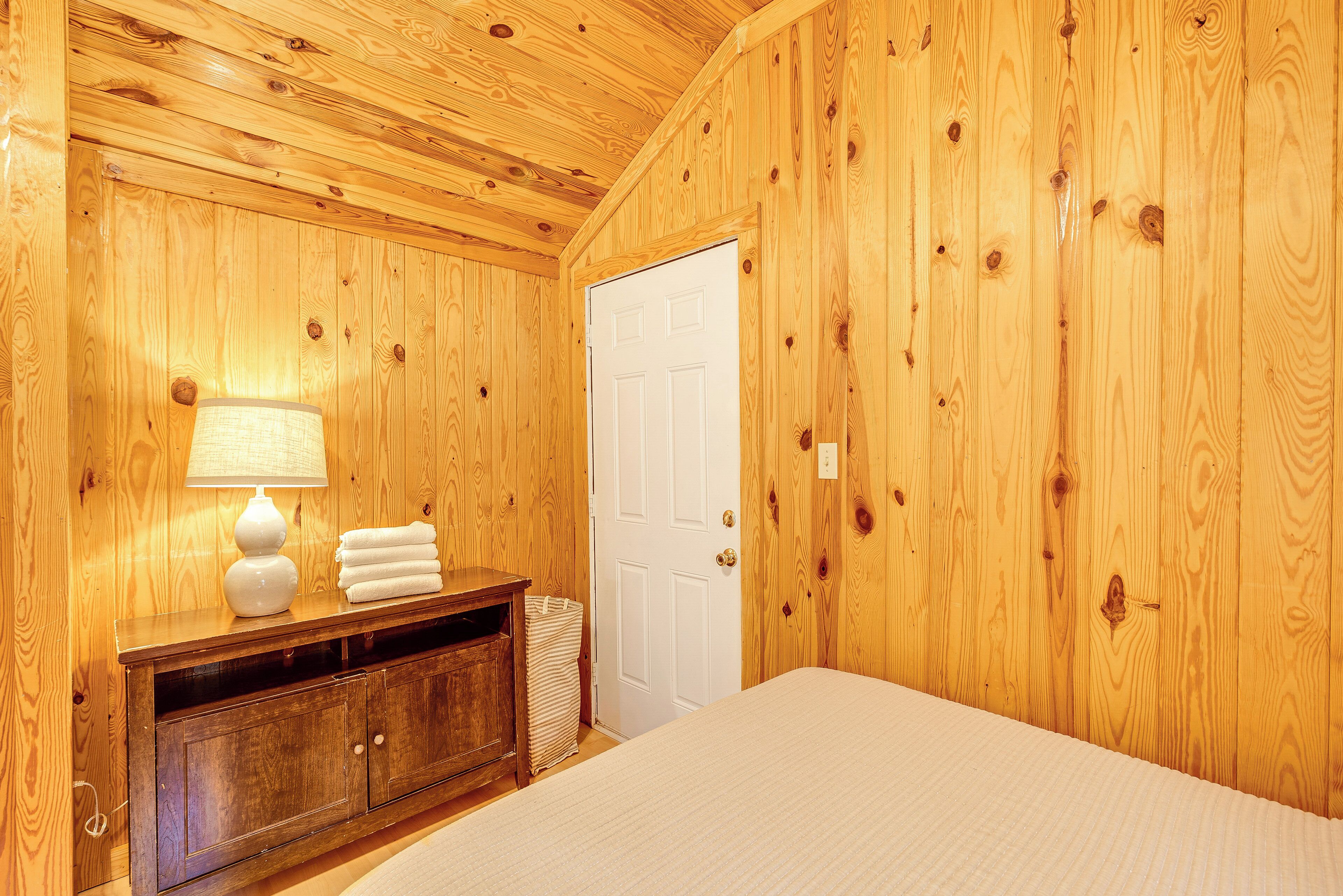 Una habitación con paneles de madera, una cama, un tocador, una lámpara y una puerta.