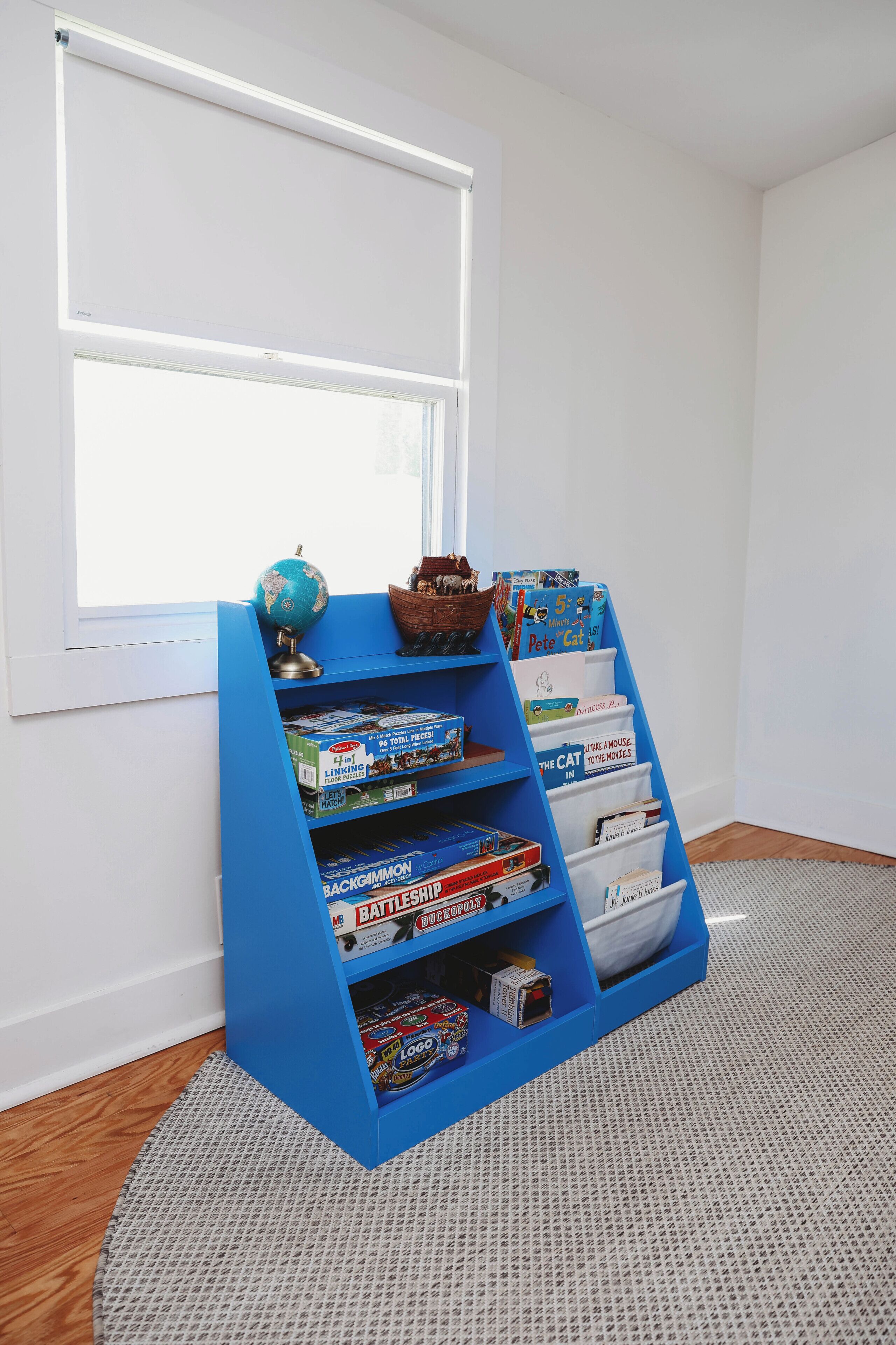 A blue bookshelf with books and toys, positioned near a window with white trim.
