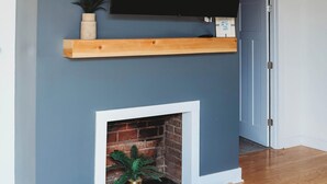 Una sala de estar con televisión montada en la pared, un estante de madera, una chimenea de ladrillo y una puerta.
