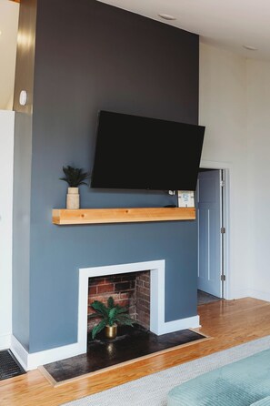 A living room with a TV mounted on a wall, a wooden shelf, a brick fireplace, and a door.