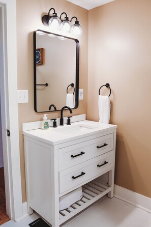 A bathroom with a white vanity, a mirror with lights, a towel ring, and a soap dispenser.
