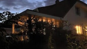 Una casa con porche cubierto y una ventana iluminada, rodeada de vegetación.