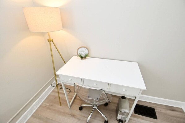 Uma mesa branca com gavetas, uma cadeira transparente, uma luminária e um pequeno relógio na parede.