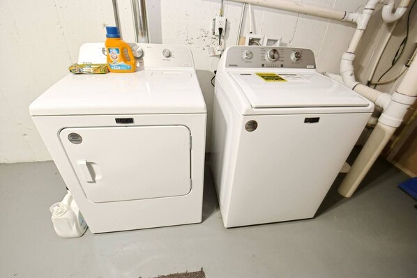 Deux machines à laver à chargement frontal, de couleur blanche, se trouvent dans une buanderie où l’on peut voir des tuyaux et une bouteille de lessive posée sur la machine de gauche.
