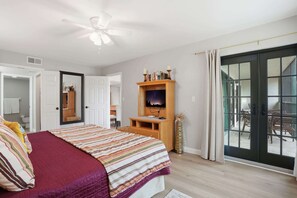A bedroom with a bed, a wooden TV stand, a ceiling fan, a sliding glass door to a balcony, and a white door leading to another room.