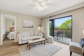 A modern living room with a sofa, a coffee table, a ceiling fan, and a view of a balcony with outdoor furniture.