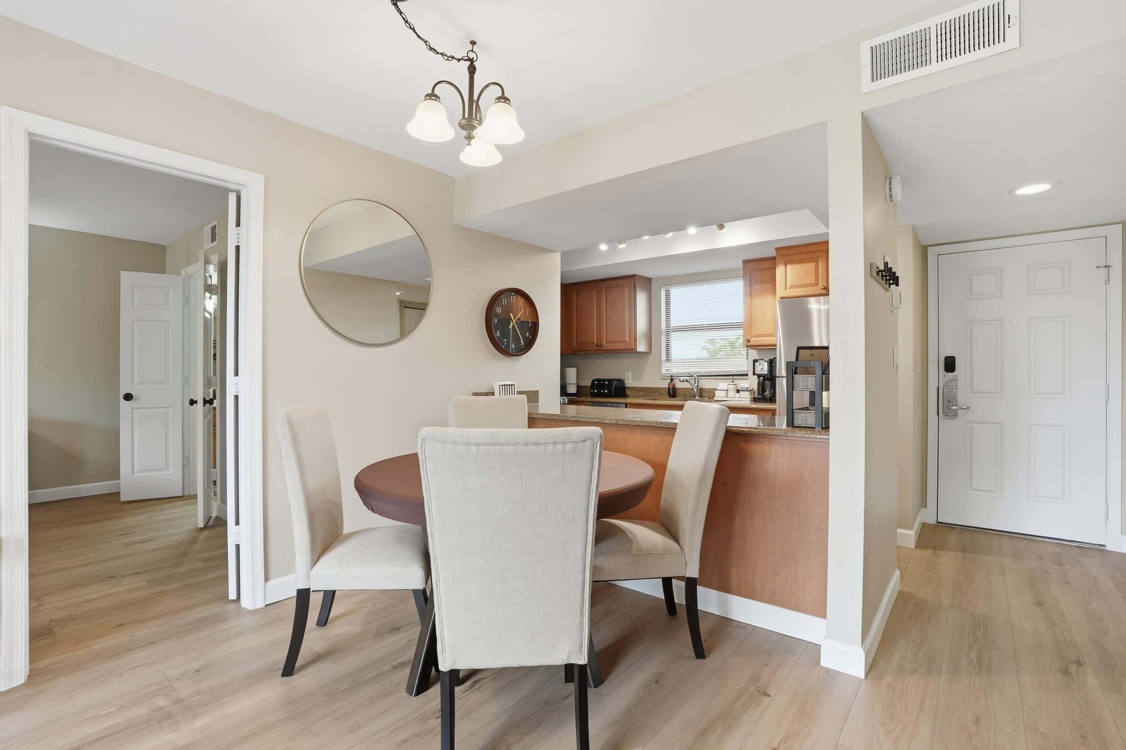 A dining area with a round table and chairs, a kitchen in the background, and a clock on the wall.