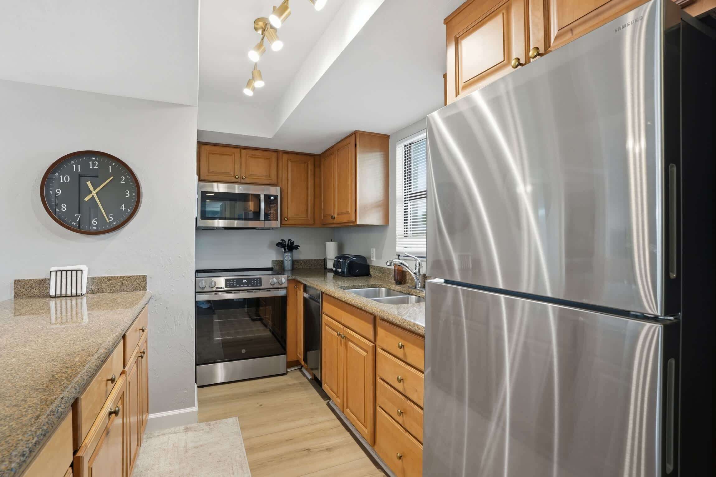A modern kitchen with stainless steel appliances, wooden cabinets, a granite countertop, and a wall clock.