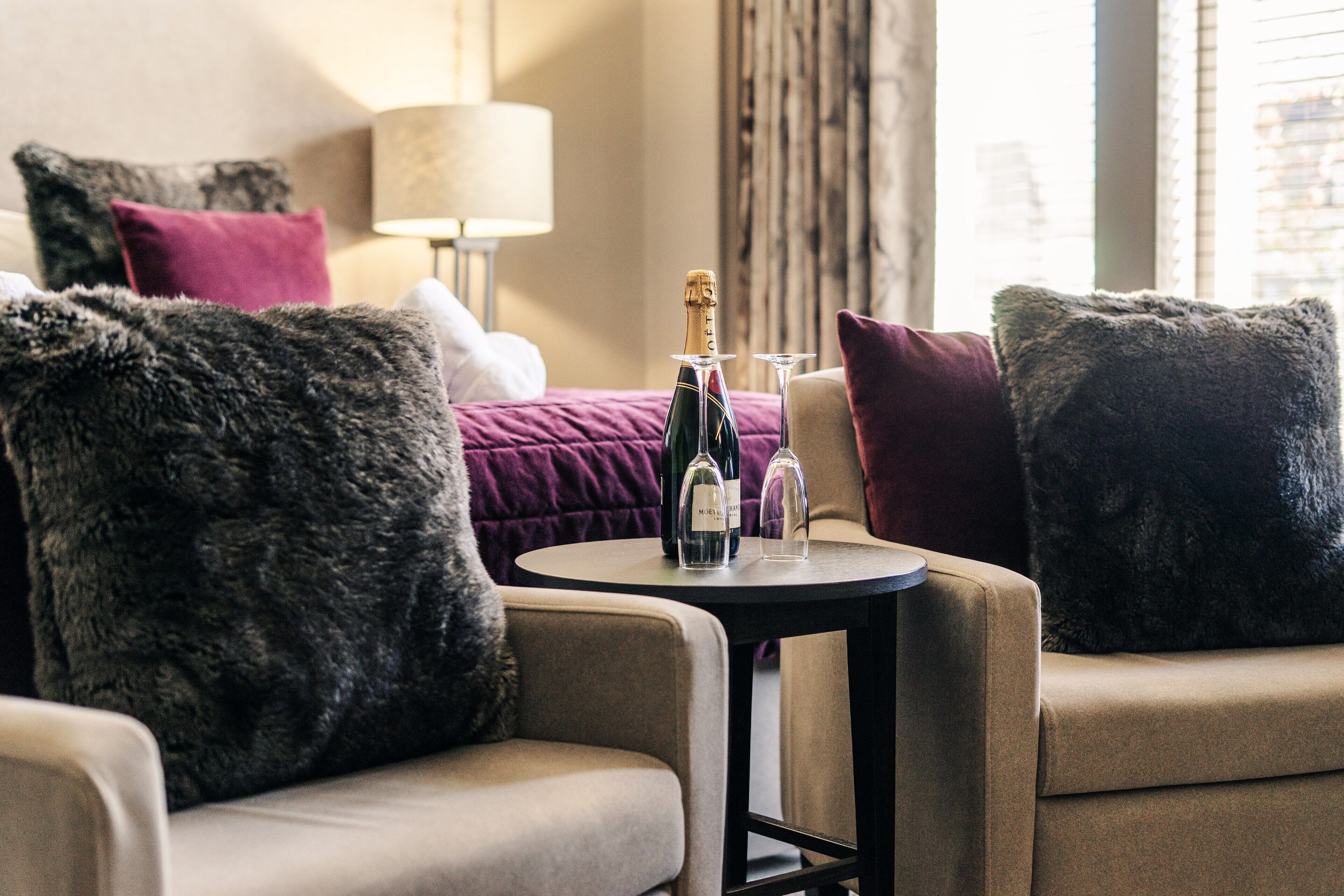 Une chambre d’hôtel avec un lit, deux fauteuils, une petite table avec une bouteille et des verres, et une lampe.