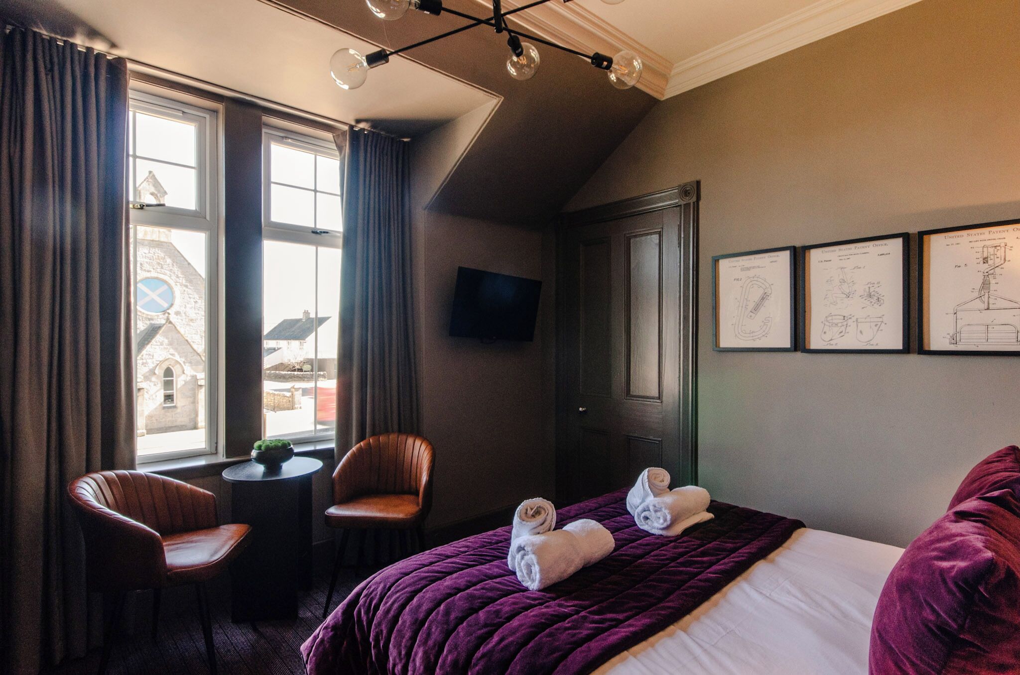 A hotel room with a bed, two chairs, a small table, a TV, and large windows with curtains.