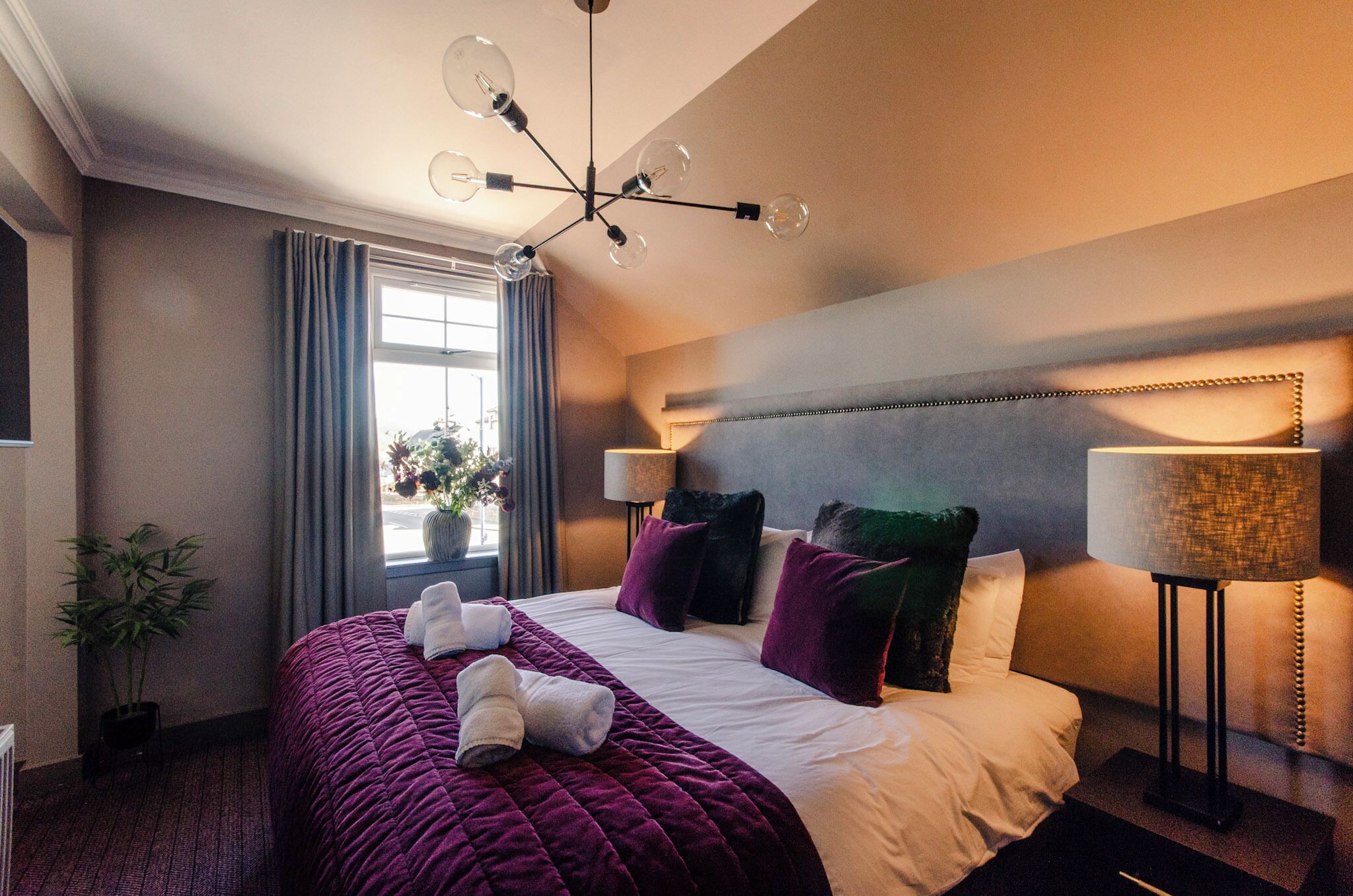 A hotel room with a large bed, a modern chandelier, a window with curtains, and a bedside lamp.