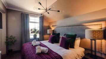 A hotel room with a large bed, a modern chandelier, a window with curtains, and a bedside lamp.