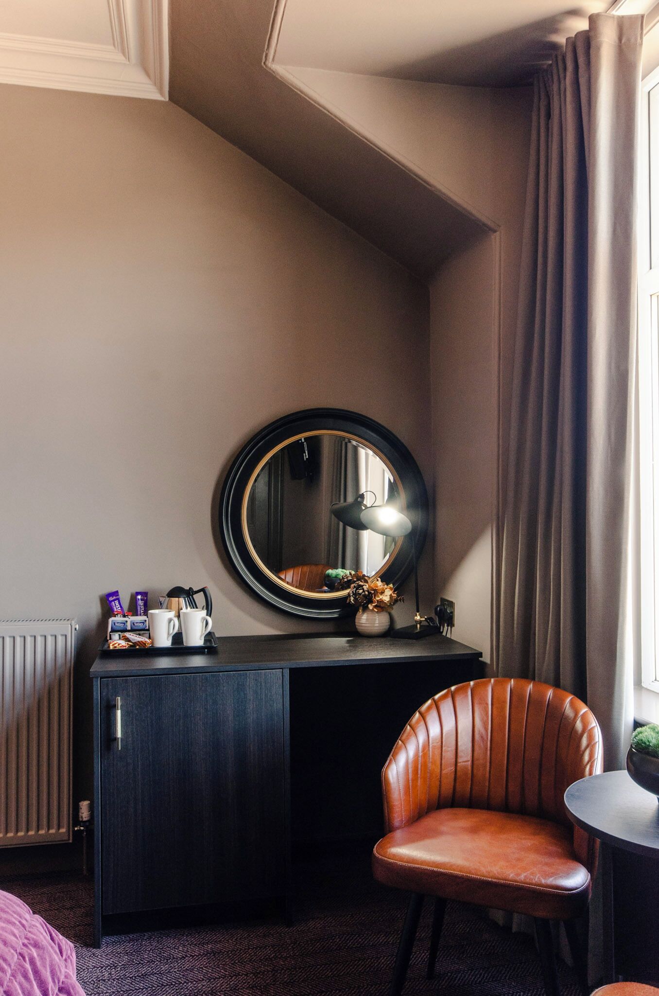 A hotel room with a dark wooden desk, a round mirror, a brown leather chair, and a small table with a lamp.