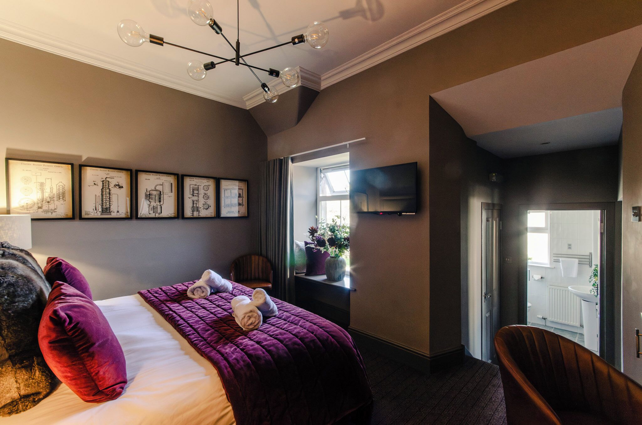 Une chambre à coucher avec un lit, une literie violette, une fenêtre avec des rideaux, un téléviseur à écran plat et des dessins d’architecture encadrés accrochés au mur.