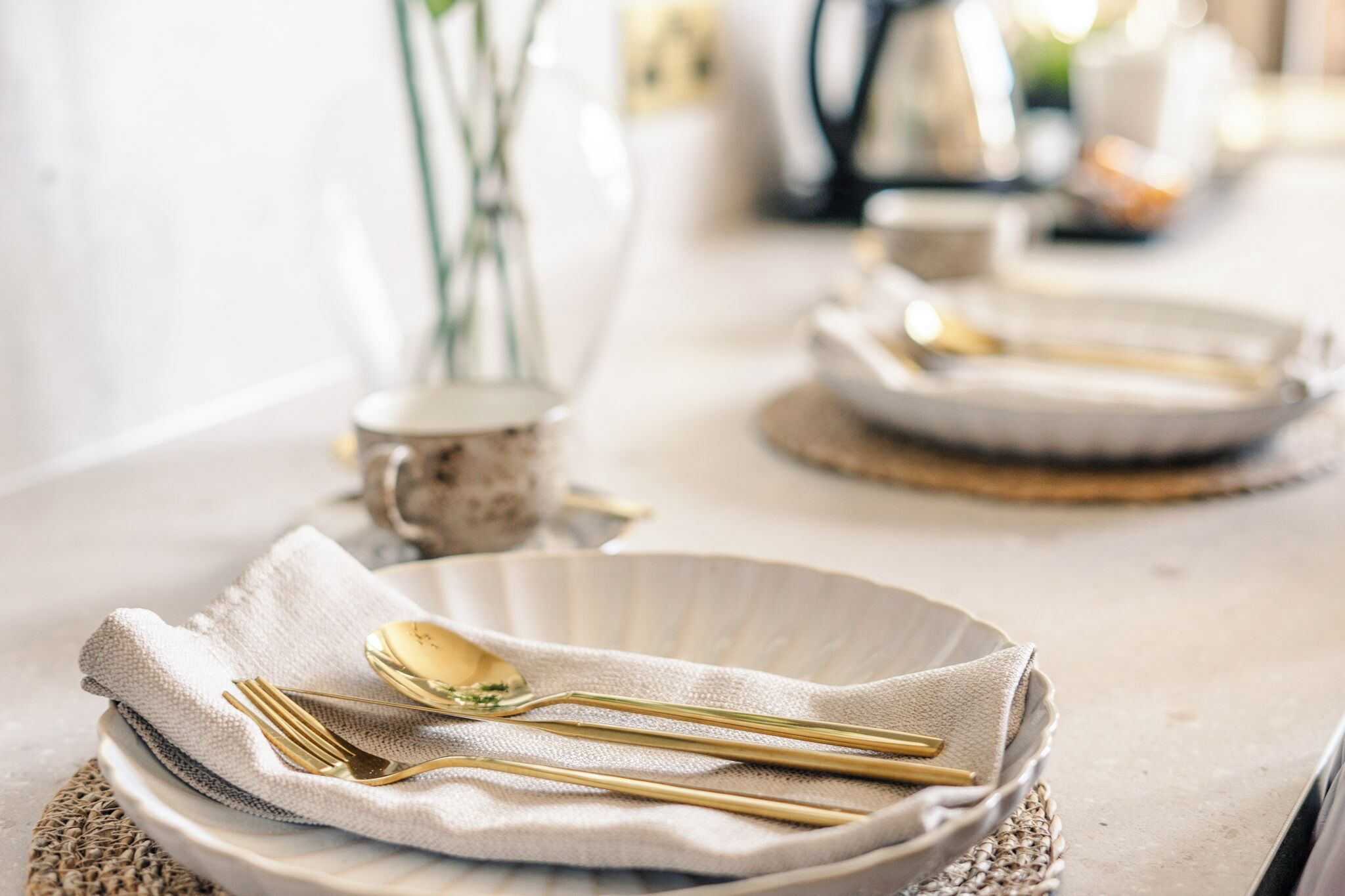 A table set with gold cutlery and a white napkin, with a vase and a teapot in the background.