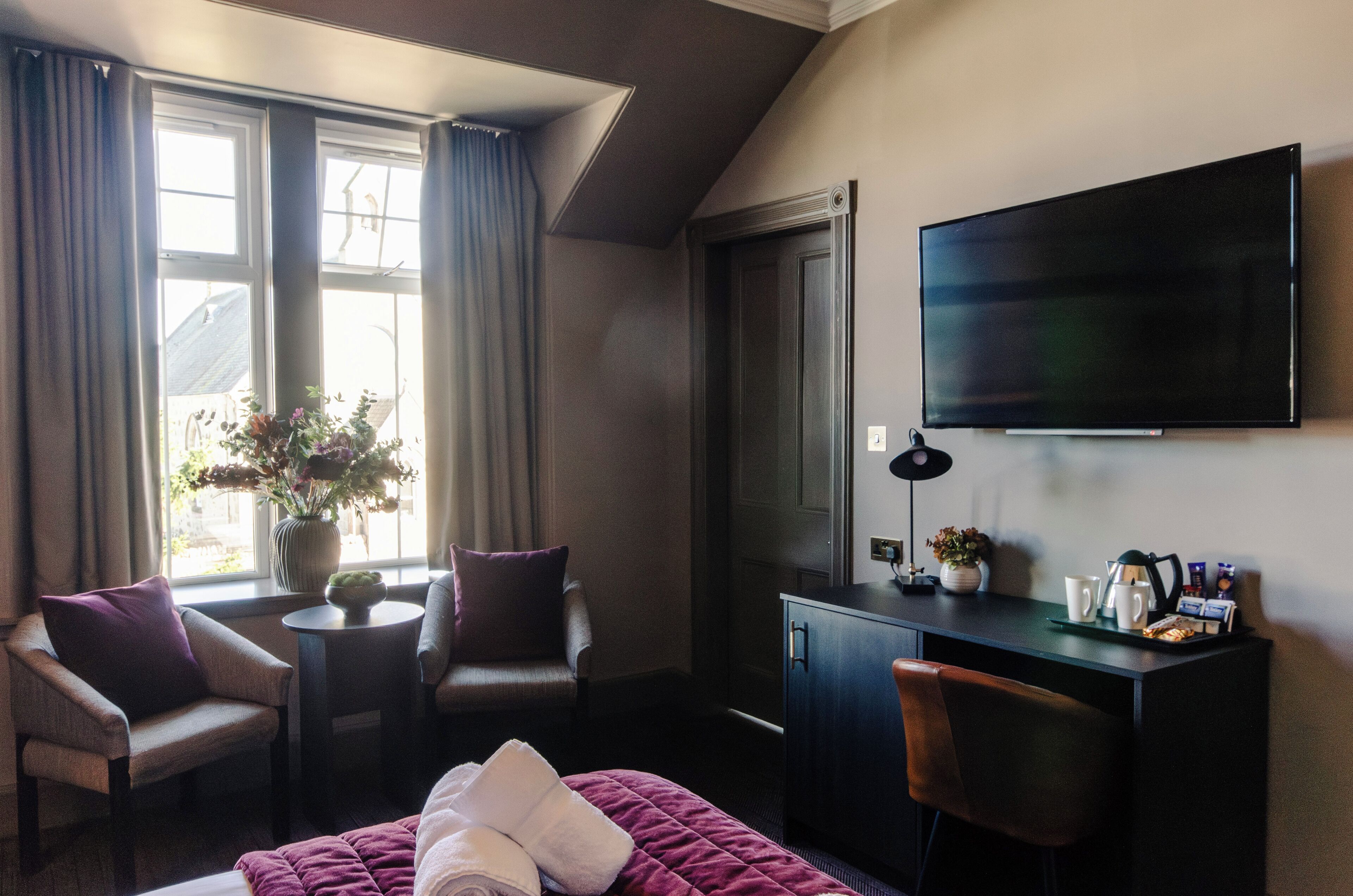 A hotel room with a bed, two armchairs, a desk, a flat-screen TV, a window with curtains, and a vase with flowers.