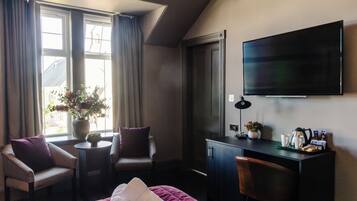 A hotel room with a bed, two armchairs, a desk, a flat-screen TV, a window with curtains, and a vase with flowers.