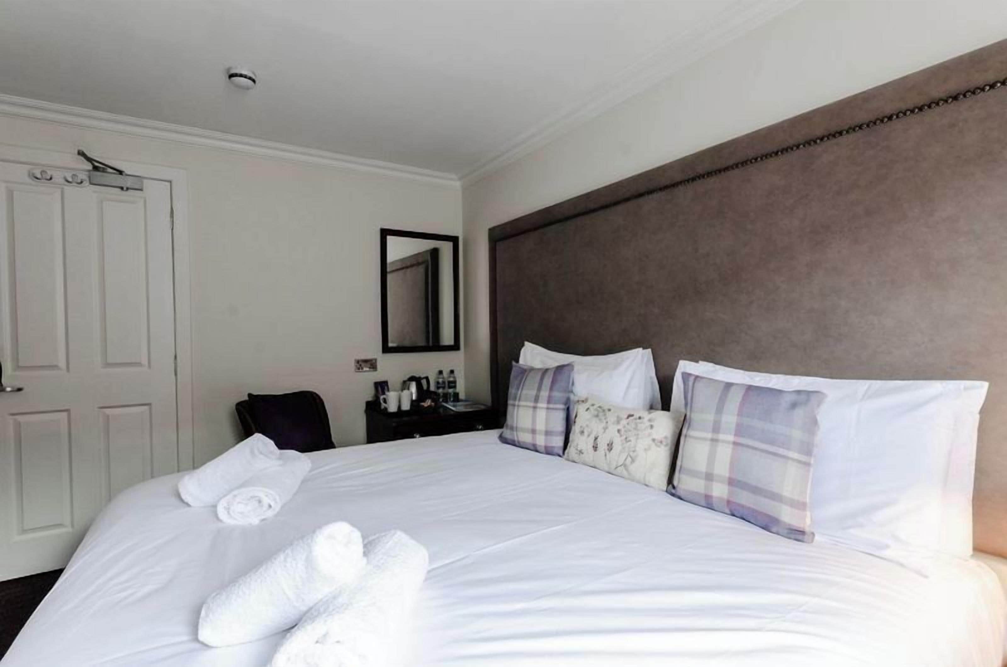 A neatly made bed with white linens and a set of white towels, a dark headboard, and a small bedside table with a mirror and toiletries.