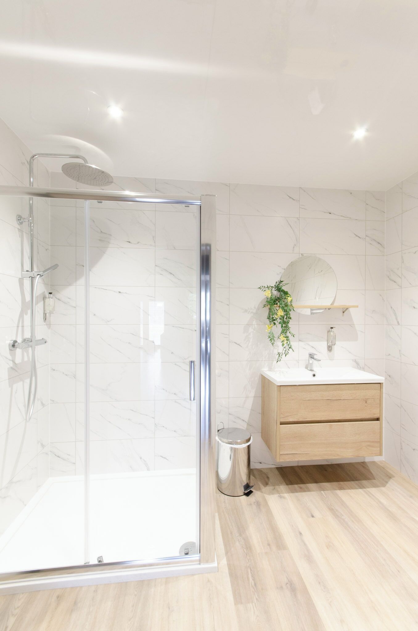 A modern bathroom with a glass-enclosed shower, a wooden vanity with a mirror, and a small trash bin.