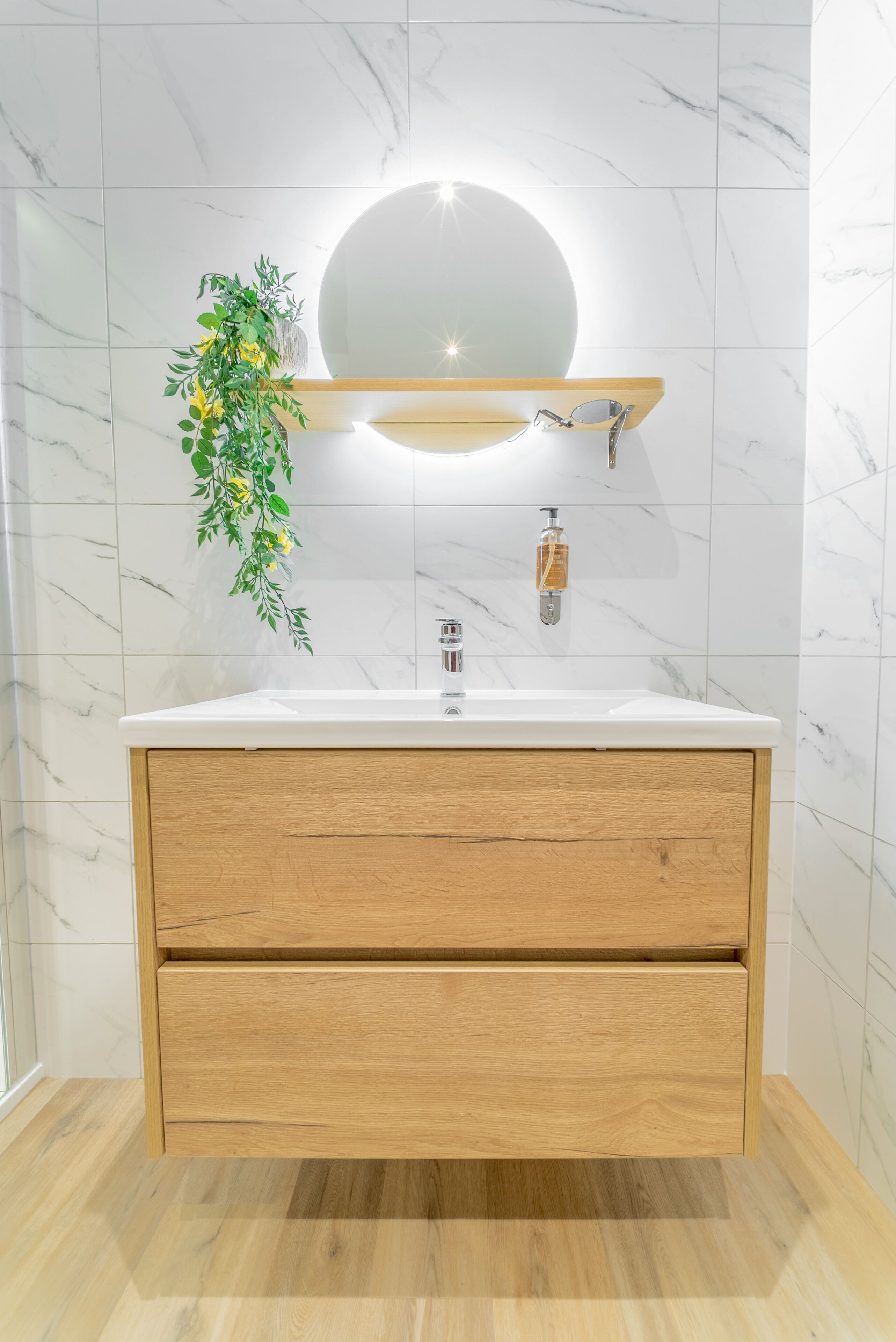 Un baño moderno con pared de azulejos de mármol, tocador de madera con un lavamanos y un espejo redondo encima.