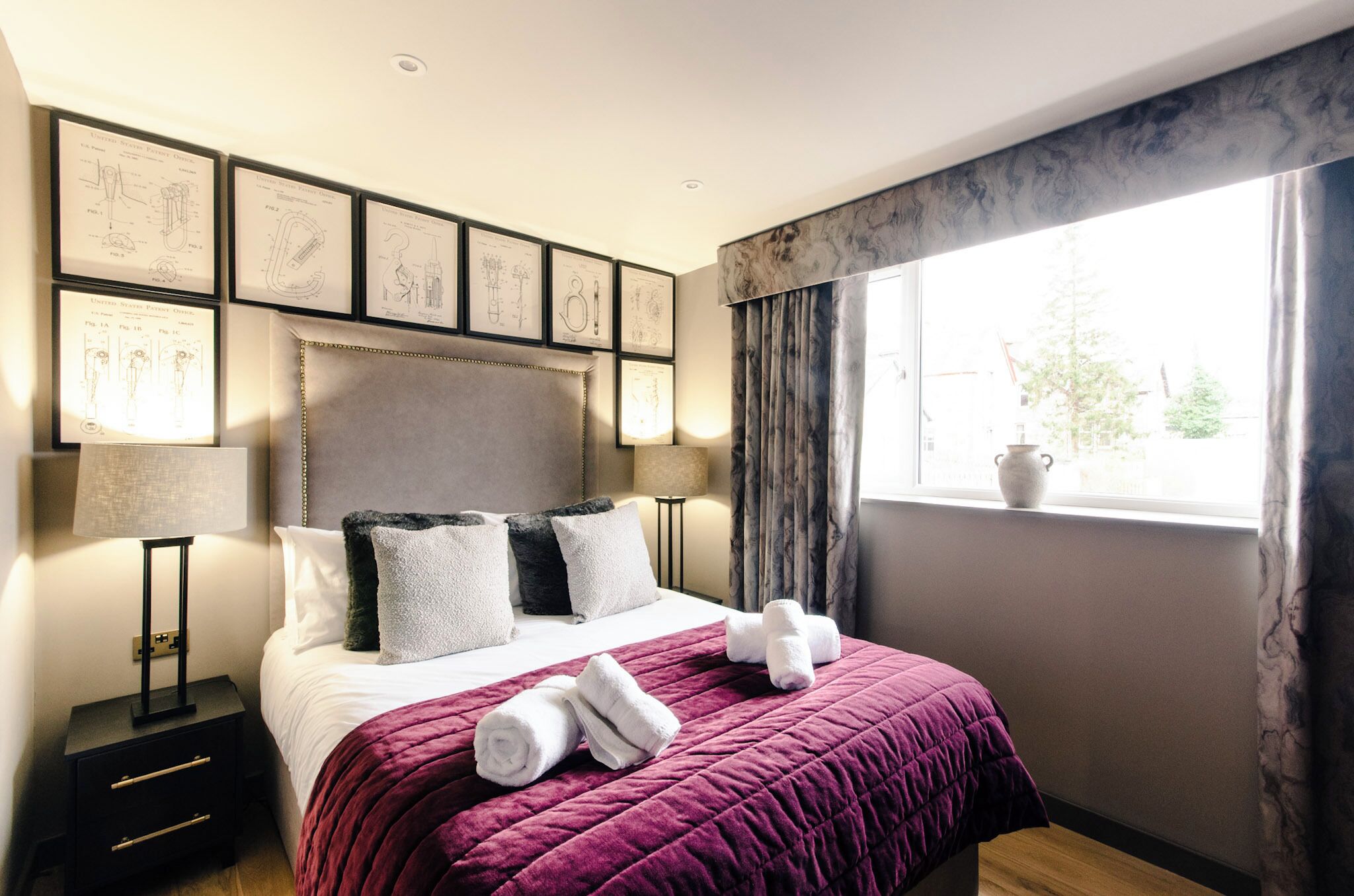 Una cama bien tendida con una colcha morada, almohadas blancas y toallas enrolladas. Hay dibujos enmarcados en la pared sobre la cama.