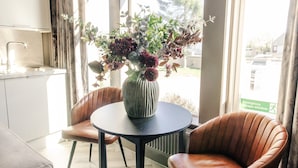 Une cuisine moderne avec un coin repas comprenant une table ronde et deux chaises, un vase avec des fleurs et de grandes fenêtres avec des rideaux.