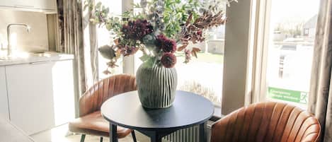 Cocina moderna con comedor que incluye una mesa redonda y dos sillas, un florero con flores y ventanas amplias con cortinas.