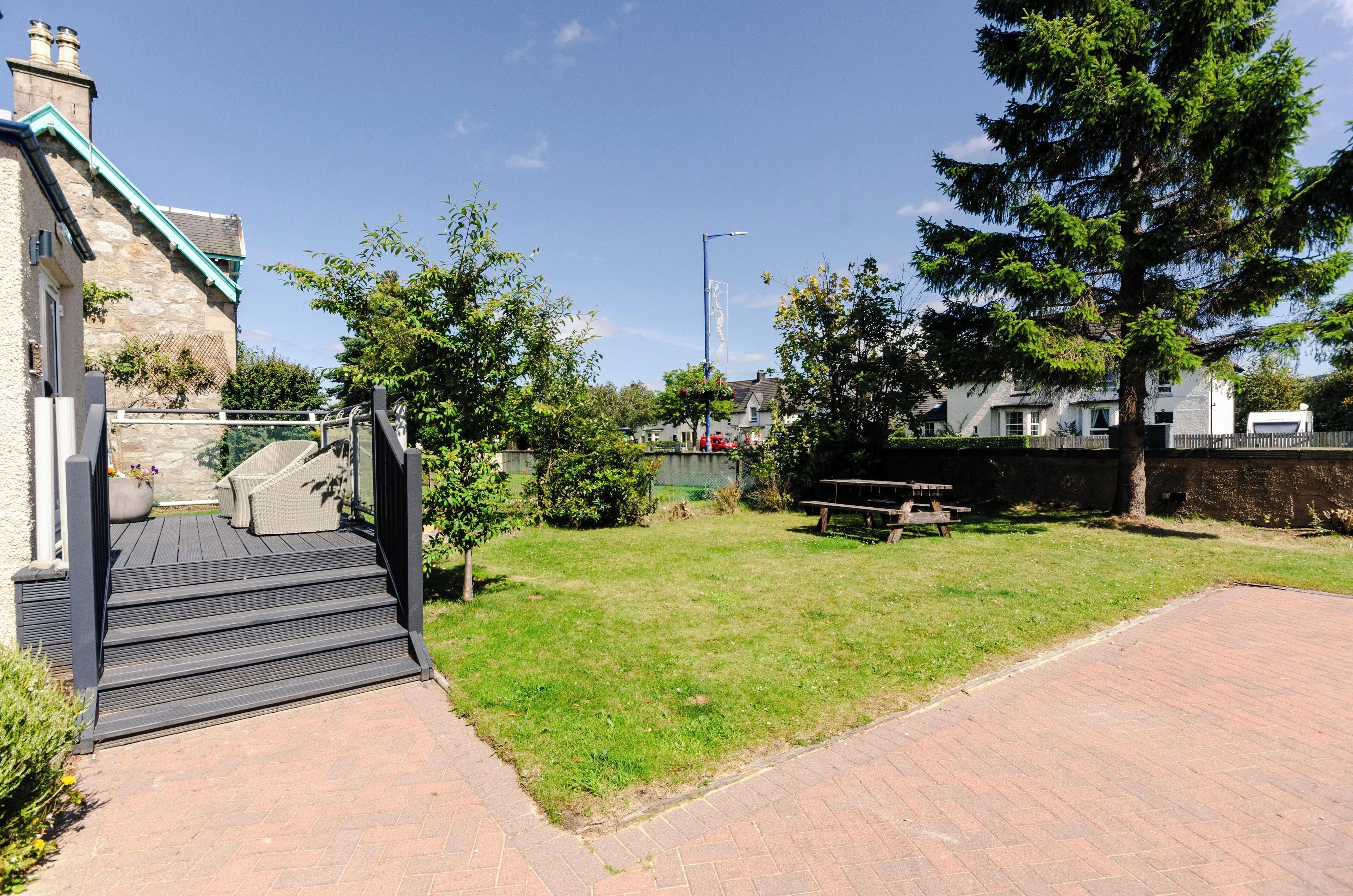A paved pathway leading to a wooden deck with a bench, surrounded by green grass and trees.