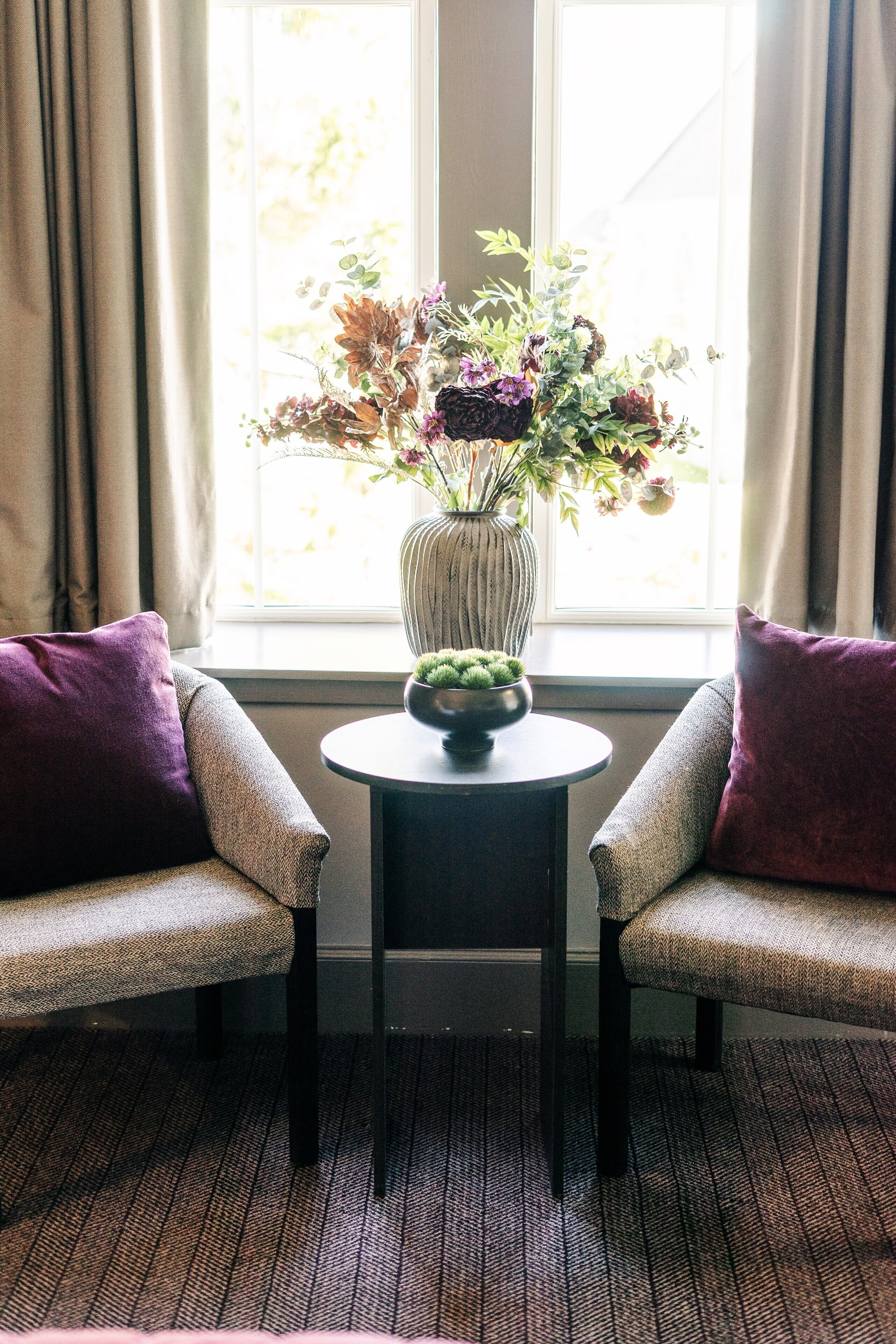 A room with two armchairs, a small table with a vase of flowers, and a window with curtains.