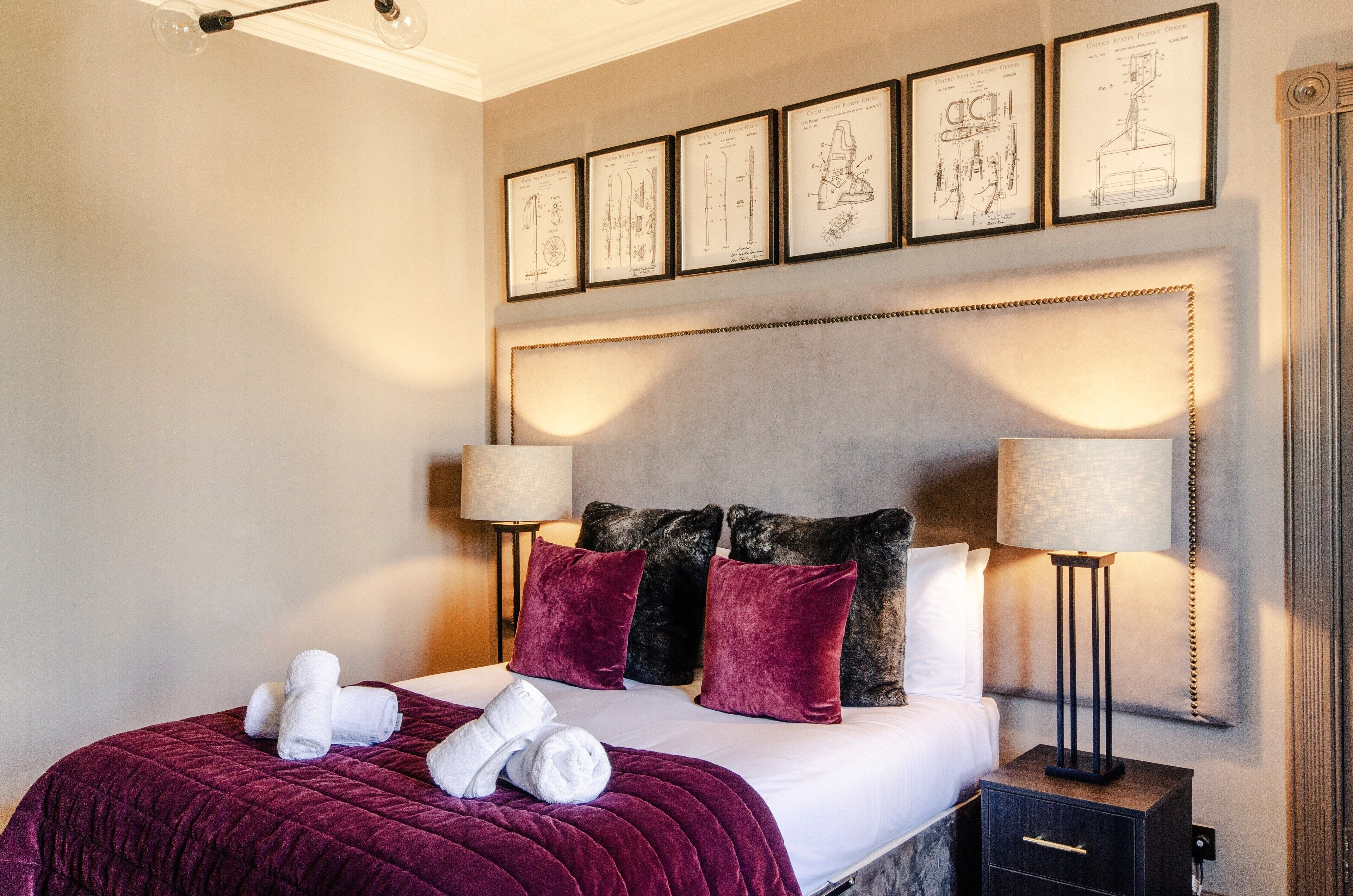 Habitación de hotel con una cama grande, dos lámparas de noche, cabecera con dibujos arquitectónicos enmarcados y mesita de noche con cajón.