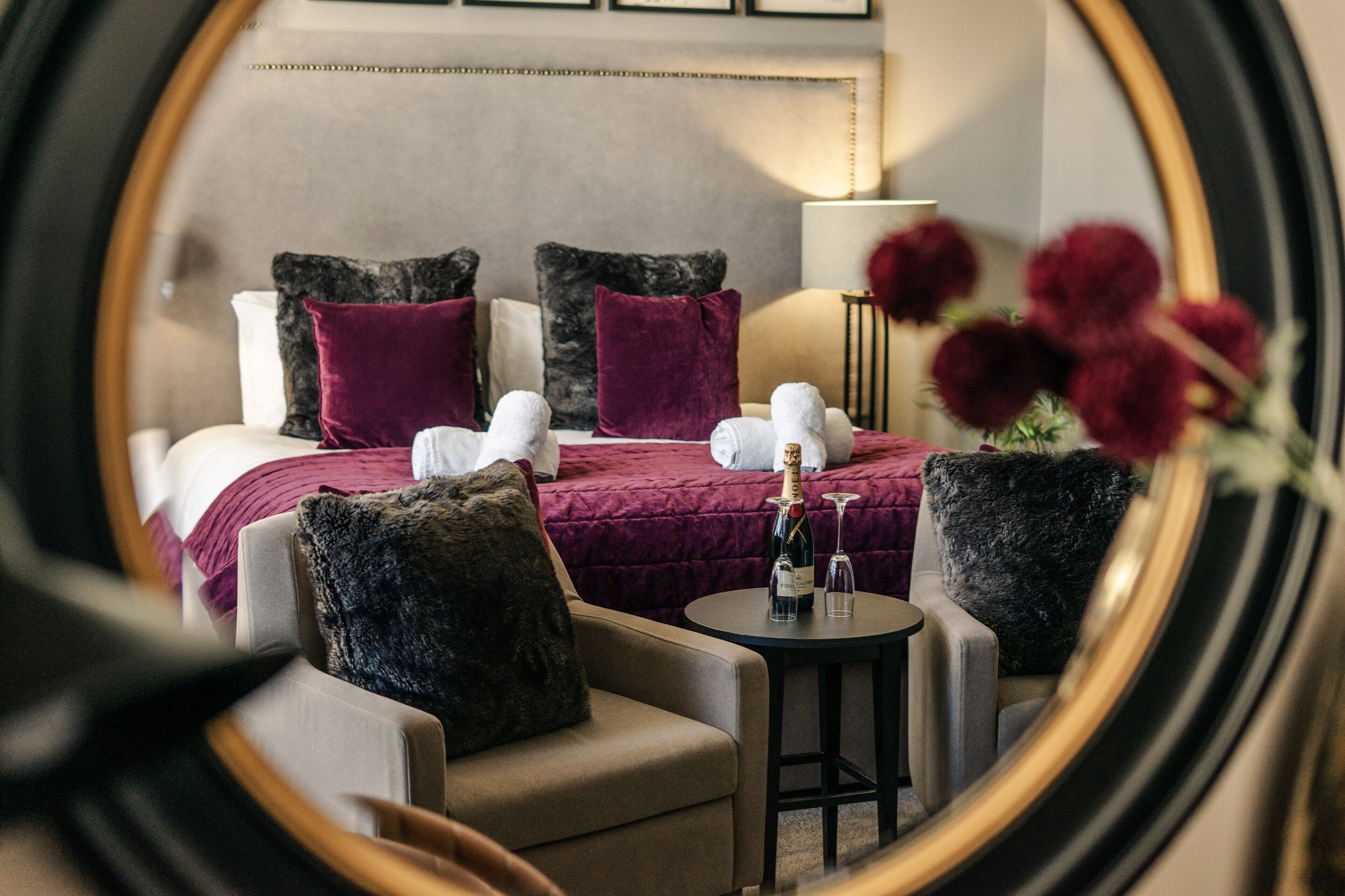 A hotel room with a bed, two armchairs, a small table with a bottle and glasses, and a mirror reflecting the room.