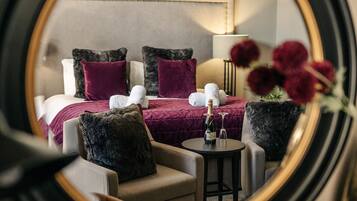 A hotel room with a bed, two armchairs, a small table with a bottle and glasses, and a mirror reflecting the room.